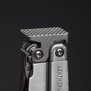 May include: Close-up of a multi-tool with a textured, silver surface. The tool has multiple components, including a textured top and the word "LEATHERMAN" printed on the side. The background is dark.