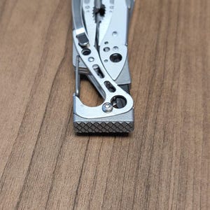 Könnte beinhalten: Ein silbernes Multitool mit verschiedenen sichtbaren Komponenten, darunter eine Zange, ein Messer und ein Karabinerhaken. Das Werkzeug besteht aus Metall und hat einen strukturierten Griff am Ende. Der Hintergrund ist eine Holzoberfläche.