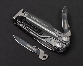 May include: A silver Leatherman Surge multi-tool with a partially open blade and an additional blade lying next to it. The multi-tool has the word "SURGE" printed on it. The tools are on a black surface.