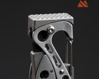May include: Close-up of a multi-tool with a textured, rectangular top and a carabiner clip. The tool is made of brushed metal and features various cut-out shapes and a small screw. The background is a solid, dark gray.