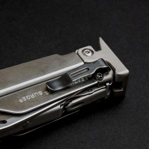 Può includere: Un utensile multifunzione argentato con finitura in metallo spazzolato e la parola SURGE incisa sul lato. L'utensile include una clip da tasca in metallo nero fissata con una piccola vite, un cacciavite a testa piatta e un'estremità a forma di T.