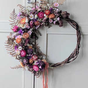 May include: A half-moon wreath composed of dried flowers in pink, orange, purple, and white hues. The floral arrangement is set on a natural grapevine base, with dried ferns adding texture. A peach ribbon is tied at the base of the flowers.