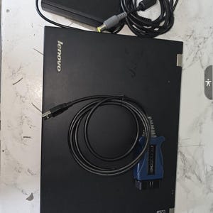 May include: Black Lenovo ThinkPad laptop with a power adapter and a black cable. A blue and black diagnostic tool with the word "MONGOOSE" is also present. The laptop is on a light-colored surface.