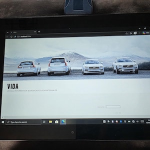 May include: A black tablet displaying the VIDA Vehicle Information and Diagnostics for Aftersales web interface. The screen shows a banner with four white Volvo cars against a mountain landscape, a login field, and a Windows taskbar at the bottom.
