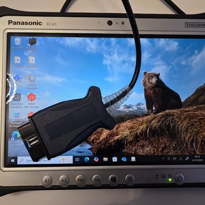 Könnte beinhalten: Panasonic Toughpad FZ-G1 Tablet mit einem schwarzen Diagnoseanschluss. Der Bildschirm zeigt einen Bären in einer Berglandschaft. Das Tablet hat einen silbernen Rahmen und schwarze Zierleisten, mit Funktionstasten unten.