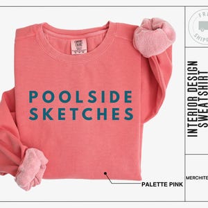 Puede incluir: Una sudadera de color rosa coral con el texto "POOLSIDE SKETCHES" en letras azul turquesa. La sudadera está doblada y se muestra sobre un fondo blanco.