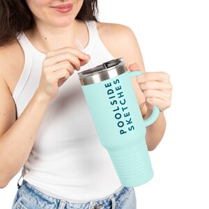 Puede incluir: Un vaso térmico azul claro con asa y tapa plateada. El vaso tiene las palabras "Poolside Sketches" impresas verticalmente en azul oscuro. Una pajita transparente está insertada en la tapa.