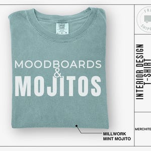 May include: A light blue t-shirt with the text "MOODBOARDS & MOJITOS" printed in white. The text "MILLWORK MINT MOJITO" is printed below the design. The t-shirt is folded and displayed on a white background.