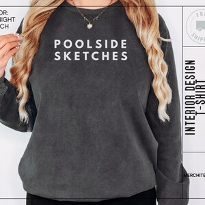 Puede incluir: Una sudadera gris oscuro con el texto "POOLSIDE SKETCHES" impreso en blanco en la parte delantera. La sudadera está siendo modelada por una persona con cabello largo y rubio. El texto "INTERIOR DESIGN T-SHIRT" está impreso en el lado derecho de la imagen.