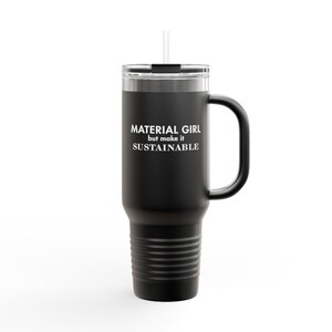 Puede incluir: Taza de viaje de acero inoxidable negro con asa y pajita. La taza tiene el texto "MATERIAL GIRL but make it SUSTAINABLE" impreso en ella.