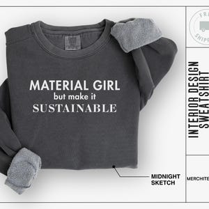 Puede incluir: Una sudadera gris oscuro con el texto "MATERIAL GIRL but make it SUSTAINABLE" impreso en blanco. La sudadera está doblada y se muestra sobre un fondo blanco.