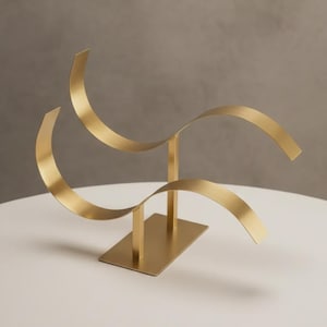 Könnte beinhalten: Eine goldfarbene Metallskulptur mit rechteckiger Basis und zwei vertikalen Stützen, die geschwungene, bandartige Elemente halten. Die Skulptur hat ein modernes, abstraktes Design und steht auf einer weißen Oberfläche.