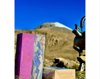 Könnte beinhalten: Zwei Schachteln, eine lila und eine beige, liegen auf Gras mit einem Berg im Hintergrund und einem klaren blauen Himmel. Die lila Schachtel hat ein dekoratives Design mit Text. Rechts befindet sich ein schwarzer Wasserkocher.