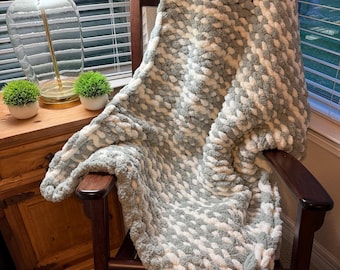 May include: A chunky knit blanket in a rocking chair. The blanket is a mix of gray and white, with a textured, woven appearance. The chair is dark wood, and the blanket is draped over the back and seat.