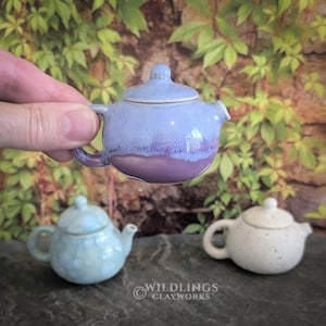 May include: A small, ceramic teapot with a lid, held by a hand. The teapot is primarily light blue with a purple base. Two other miniature teapots, one light blue with white spots and one white with speckles, are also visible.
