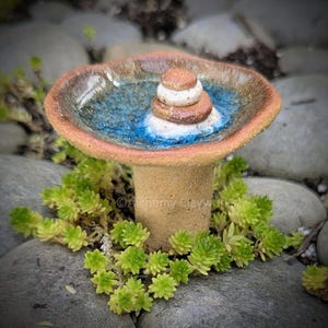 Op de afbeelding: Een handgemaakte keramische vogelbad in de vorm van een paddenstoel. De kom heeft een blauw en bruin geglazuurd interieur met opgestapelde stenen. De basis is lichtbruin, omringd door groene planten en geplaatst op een bed van stenen.