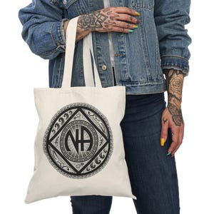 Puede incluir: Una bolsa de lona blanca con un diseño gráfico en blanco y negro de un círculo con las letras "NA" en su interior. El círculo está rodeado de olas y otros elementos decorativos.