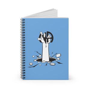 Puede incluir: Una libreta espiral azul con una ilustración en blanco y negro de una mano que sale de una grieta en el suelo. La mano sostiene un círculo negro con las letras "NA" en su interior.