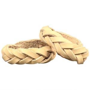 May include: Two braided bracelets crafted from natural, light beige leather. Each bracelet features a thick, three-strand plait design with a smooth, matte finish. The leather shows a visible grain texture, and the ends are secured together.