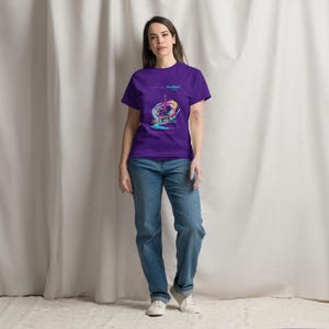 May include: A purple t-shirt with a colorful graphic design and the text "Bonjour!". The shirt is paired with blue jeans and white sneakers. The model is standing in front of a white curtain.