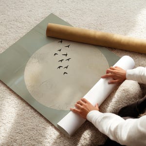 May include: A large print with a moon and birds design, with a light green border. A rolled-up print and a white rolled-up paper are next to the print. The print is on a light-colored rug.