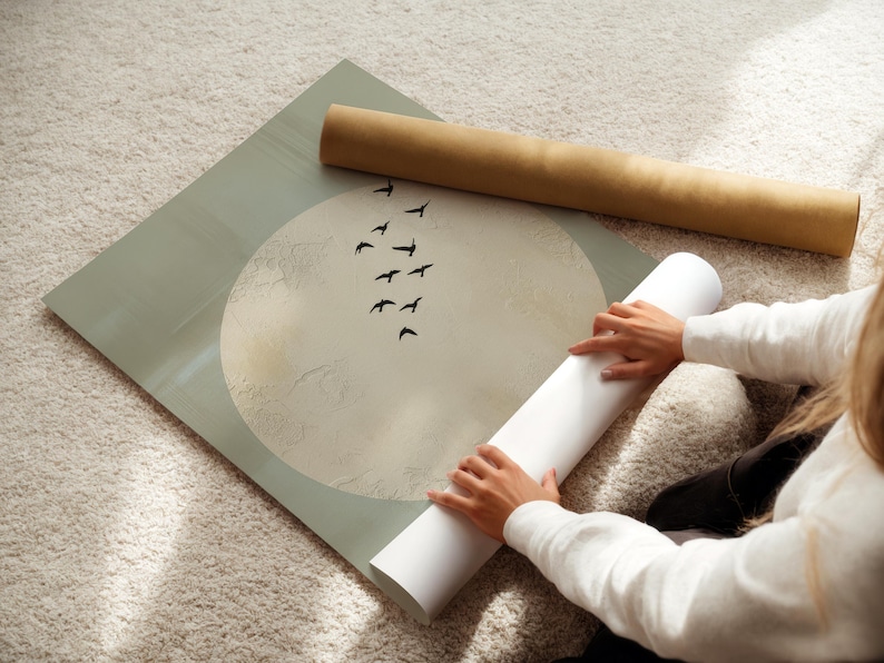May include: A large print with a moon and birds design, with a light green border. A rolled-up print and a white rolled-up paper are next to the print. The print is on a light-colored rug.