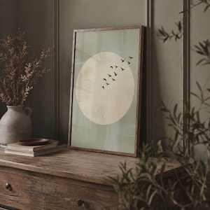 May include: A framed art print featuring a large, textured, off-white circle against a pale green background. A flock of black birds flies across the circle. The frame is a warm brown color. The artwork is displayed on a wooden surface.
