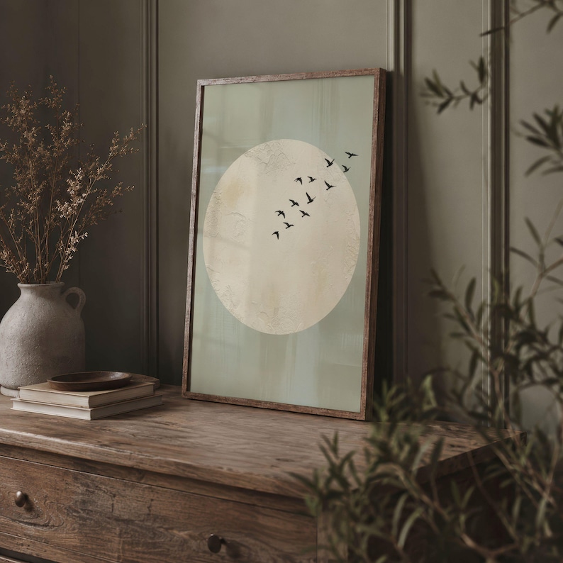 May include: A framed art print featuring a large, textured, off-white circle against a pale green background. A flock of black birds flies across the circle. The frame is a warm brown color. The artwork is displayed on a wooden surface.