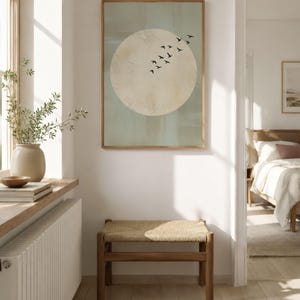 May include: A framed wall art print featuring a large, off-white circle against a pale green background, with a flock of black birds in flight. A wooden bench with a woven seat sits below the artwork. The room has a neutral color palette.