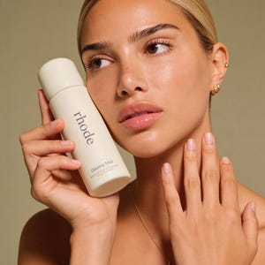 May include: A woman holds a white spray bottle labelled "rhode Glazing Mist." The bottle is cylindrical with a rounded top. The woman has blonde hair, gold earrings, and a gold necklace. The background is a solid, muted green.