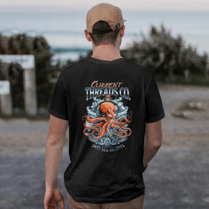 Op de afbeelding: Zwart T-shirt met een afbeelding van een octopus in een fles met de tekst "Current Threads Co." en "Deep Sea Secrets".