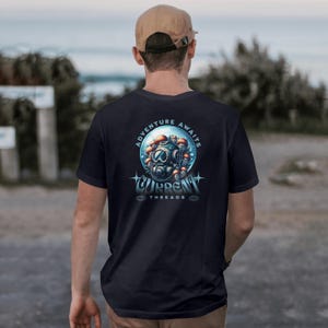 Op de afbeelding: Marineblauw T-shirt met een afbeelding op de achterkant van een duikhelm en paddenstoelen, met de tekst "ADVENTURE AWAITS" en "CURRENT THREADS". De persoon draagt een beige baseballpet.