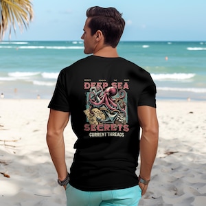 Op de afbeelding: Zwart T-shirt met een afbeelding van een octopus, anker en schatkaarten. De tekst luidt "Secrets Beneath The Waves Deep Sea Secrets Current Threads".