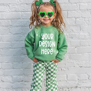 May include: A young child wearing a green sweatshirt with the text "YOUR DESIGN HERE", green heart-shaped sunglasses, and green and white checkered pants. The child is smiling and wearing green hair accessories.