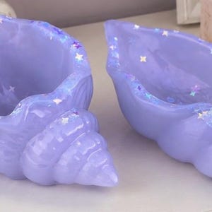 May include: Two glossy, light purple seashell-shaped bowls crafted from a smooth resin material. Each shell features a translucent finish embedded with small, reflective holographic star-shaped glitter accents along the inner edges and rims.
