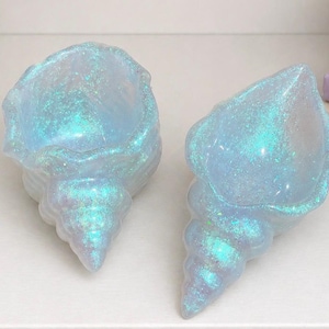 May include: Two iridescent blue seashell-shaped decorative bowls. The bowls have a glittery finish, with shades of blue and green. The shells are placed on a white surface, with a book in the background.