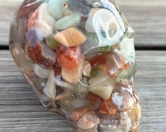 Pode incluir: Uma caveira de resina transparente preenchida com uma variedade de pedras coloridas. As pedras são em tons de branco, laranja, vermelho e verde. A caveira está sobre uma superfície de madeira.