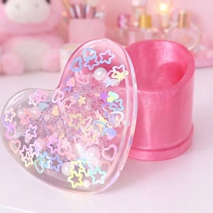 Può includere: Un portagioie rosa a forma di cuore con un coperchio trasparente pieno di stelle, cuori e perle iridescenti. La scatola è aperta, rivelando una base rosa abbinata. Lo sfondo presenta altri oggetti rosa.