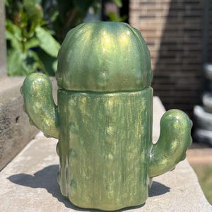 May include: A cactus-shaped container with a removable lid, crafted from a translucent, shimmering light green resin material. The object features a cylindrical body with two side arms and a rounded top, all adorned with small, raised bumps.
