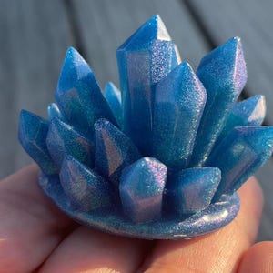 May include: A cluster of blue crystal formations with iridescent shimmer. The crystals are arranged in a radial pattern, with sharp, pointed tips. The base is a rounded shape, and the overall effect is a sparkling, gem-like appearance.