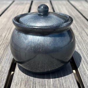 Puede incluir: Un pequeño tarro redondo de cerámica gris oscuro con tapa. El tarro tiene un acabado brillante y moteado y un pequeño pomo redondo en la parte superior de la tapa. El tarro está sobre una superficie de madera.