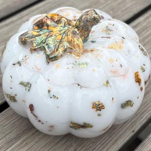 May include: A white pumpkin-shaped ornament with gold leaf detailing. The pumpkin features a stem and gold leaf accents. The surface is smooth and glossy, with gold and green flecks. A seasonal home decor item.