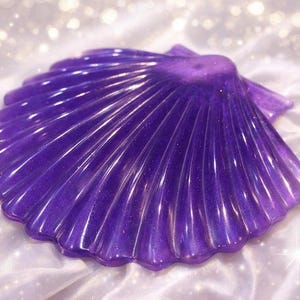 May include: A translucent, purple seashell-shaped object with a glossy finish. The shell has a ribbed texture and is set against a white, silky background with sparkling accents. The object is likely a decorative item.