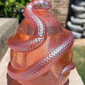 May include: A translucent orange egg-shaped container featuring a coiled snake wrapped around its exterior. The snake has an iridescent, shimmering finish that shifts between blue and purple tones, while the orange base contains fine glitter.