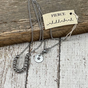 Puede incluir: Un collar de plata con un colgante en forma de herradura hecho de pequeñas cuentas de plata. El colgante está unido a una cadena de plata con un cierre de langosta. Un pequeño amuleto de plata con un recorte de estrella cuelga de la cadena.