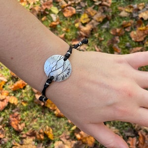 Könnte beinhalten: Ein Armband aus schwarzem Band mit einem runden Silberanhänger, der ein Bergkettenmotiv und das Wort "Home" zeigt.