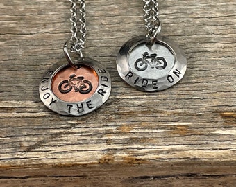 May include: Two silver necklaces with round pendants. One pendant is copper and says "Enjoy the Ride" with a bicycle image. The other pendant is silver and says "Ride On" with a bicycle image.