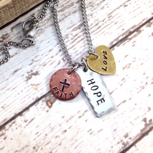 May include: A silver chain necklace with three charms: a copper charm with a cross and the word "FAITH", a silver charm with the word "HOPE", and a gold heart charm with the word "LOVE".
