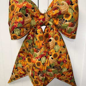 May include: A large fabric bow featuring a vibrant autumn-themed print on a black background. The pattern displays cornucopias, orange pumpkins, yellow sunflowers, and colorful fall leaves. The bow has two wide top loops and two long, pointed tails.