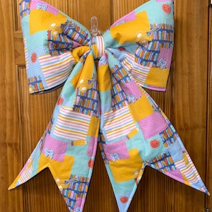 May include: A large fabric bow featuring a colorful patchwork pattern of school-themed illustrations. The design includes rows of books, yellow scissors, pencils, small blue bows, and smiling red apples on backgrounds of yellow, light blue, and pink.